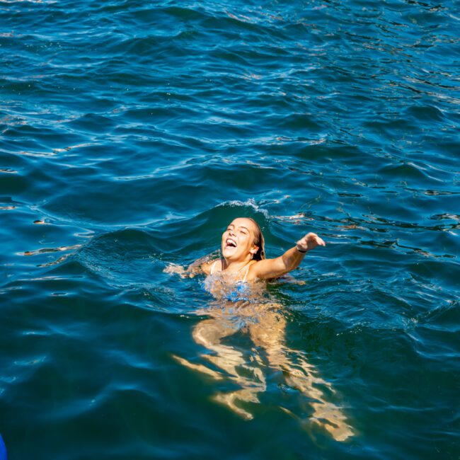 A person in a blue swimsuit is swimming and smiling in a large body of water, appearing to enjoy the swim with one arm outstretched. Other swimmers are visible in the background, and the deep blue-green water adds to the serene setting. Nearby, luxury yachts from The Yacht Social Club enhance the scene.