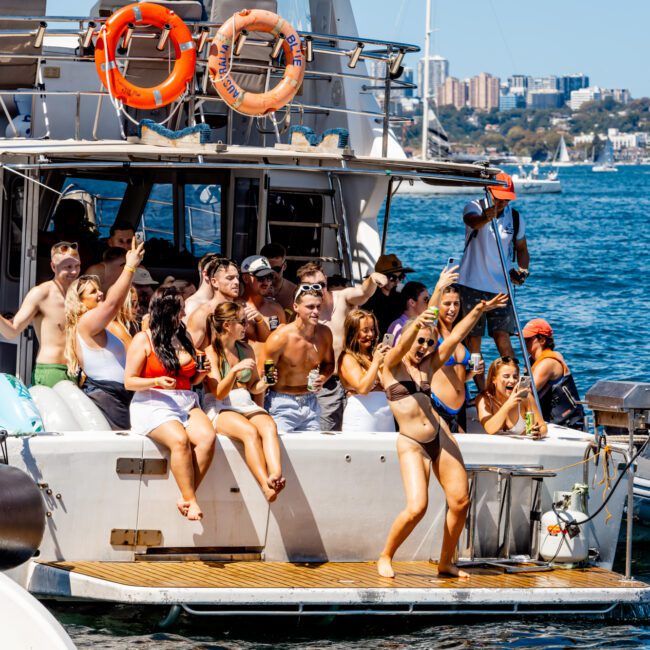 A group of people enjoying themselves on a boat from The Yacht Social Club Sydney Boat Hire. They are wearing swimsuits, taking selfies, and some are sitting on the edge of the boat. The backdrop features a large body of water with other boats and buildings in the distance. It's a sunny day.