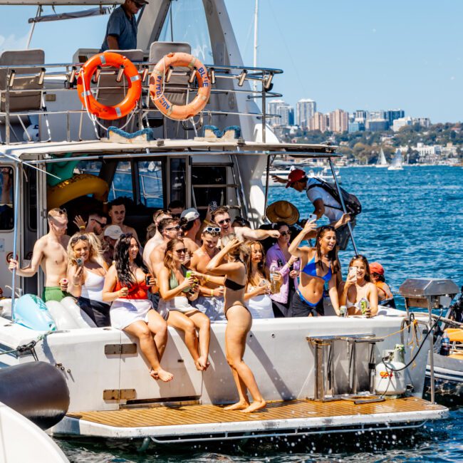 A group of people in swimwear enjoy a sunny day on a yacht, dancing and holding drinks. The yacht, chartered through Sydney Harbour Boat Hire, is anchored in a vibrant blue body of water with other boats and a cityscape visible in the background. Some people are on a smaller boat next to the yacht.