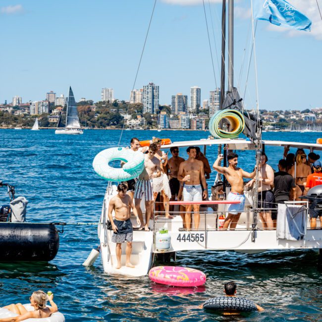 A group of people enjoy a sunny day on a sailboat with The Yacht Social Club. Some are in swimsuits on the boat, while others float nearby on inflatable tubes. The background features a city skyline with numerous buildings and other sailboats on the water.