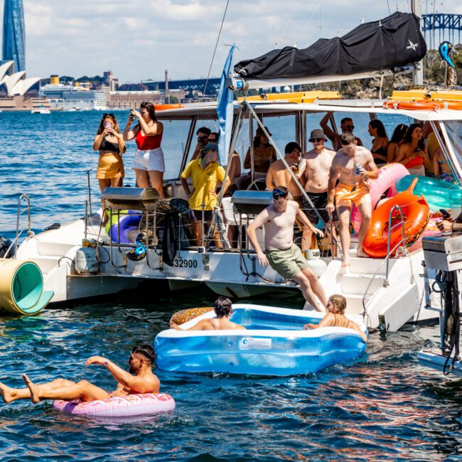 A group of people enjoying a sunny day on and around a boat in a harbor. Some are lounging on the boat, while others are in inflatable pools and tubes in the water. The Sydney Opera House and Harbour Bridge are visible in the background, showcasing Boat Parties Sydney The Yacht Social Club.