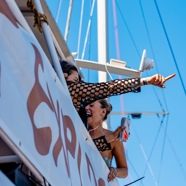 Two people are on a boat, looking cheerful. One person is pointing excitedly into the distance while the other is holding a drink and smiling. They are next to a large banner with partially visible text. The clear blue sky creates a perfect backdrop, showcasing the joy of Sydney Harbour Boat Hire with The Yacht Social Club.