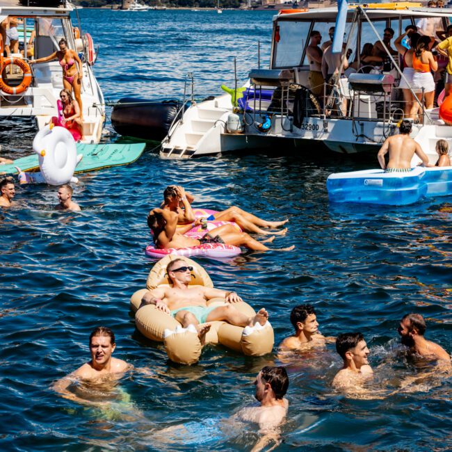 People are enjoying a sunny day on the water with floating devices and swimming, surrounded by boats. The city skyline is visible in the background, featuring tall skyscrapers and modern buildings. The atmosphere is festive and lively with multiple groups socializing at The Yacht Social Club Sydney Boat Hire.