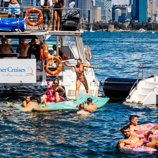 A lively scene of people enjoying time on the water in front of a city skyline. Some are on a boat, others are floating on inflatables and paddleboards. The sky is partly cloudy and the water is clear. A large modern cityscape with tall buildings is visible, typical of Boat Parties Sydney The Yacht Social Club.