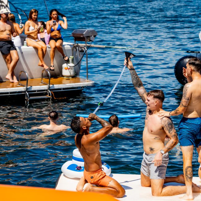 A group of people enjoying a sunny day on the water, some on boats and others swimming. Three shirtless men are on a boat, one drinking from a beer bong held by another. Several others lounge and socialize on another boat in the background, showcasing the vibrant atmosphere of Sydney Harbour Boat Hire The Yacht Social Club.