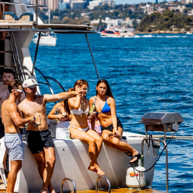 A group of people in swimwear are on the deck of a boat, smiling and enjoying a sunny day. The boat is anchored on calm blue waters, with city buildings and other boats visible in the background. Part of The Yacht Social Club, some are standing while others lounge, epitomizing Luxury Yacht Rentals Sydney.