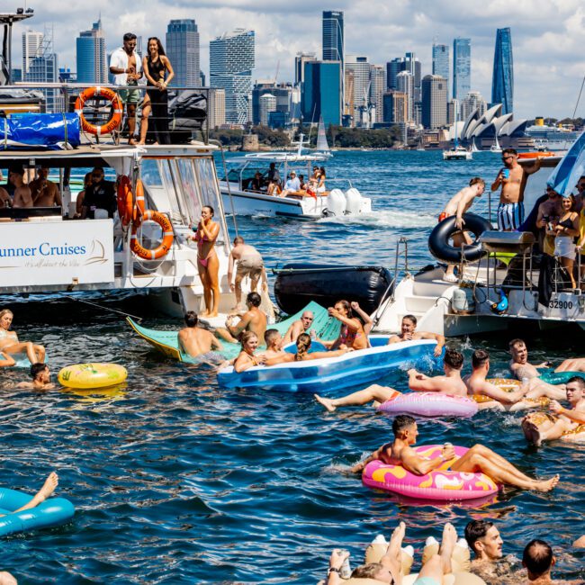 A lively scene on the water with people enjoying a sunny day on inflatable floats, boats, and yachts. The background features a city skyline under a bright blue sky with fluffy clouds. Some are swimming, while others are socializing on boats from Sydney Harbour Boat Hire The Yacht Social Club.