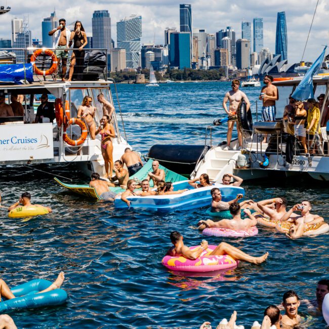 A lively boating party on a sunny day with numerous people swimming and relaxing on colorful inflatable rafts in the water. The scene includes two boats, one labeled "Rum Runner Cruises," and a city skyline in the background. Experience the festive atmosphere with Sydney Harbour Boat Hire - The Yacht Social Club.