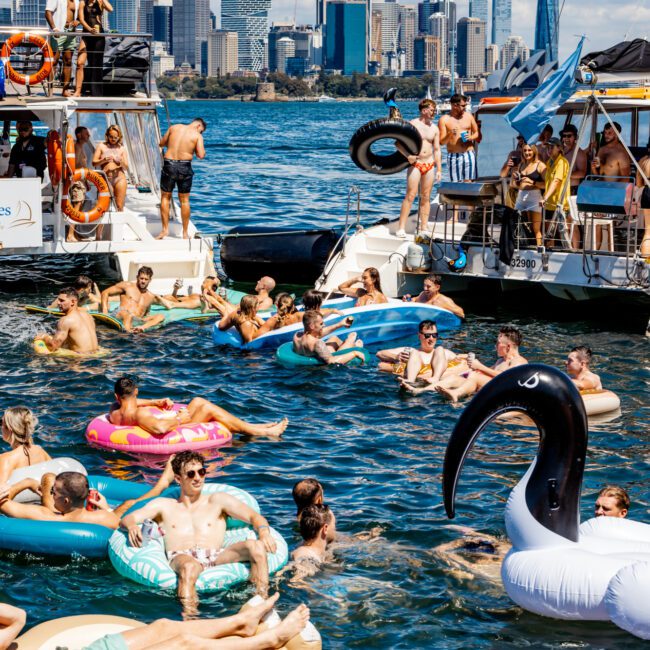 A lively scene of people enjoying a sunny day on the water with several boats nearby. Many individuals are floating on colorful inflatables, including a black and white swan. The city skyline is visible in the background under a sky with scattered clouds, all part of The Yacht Social Club Event Boat Charters.