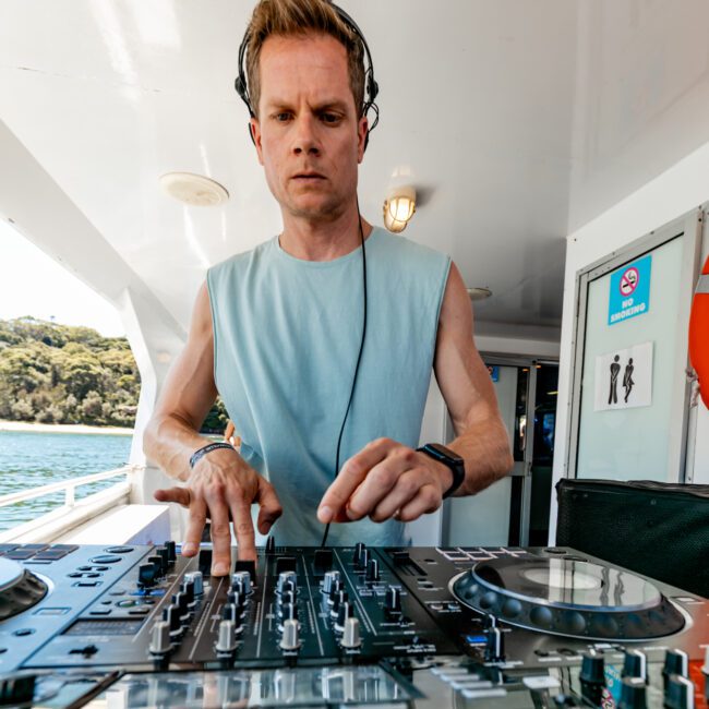 A person with short hair, wearing headphones and a sleeveless blue shirt, is focused on DJ equipment on a boat. The yacht has a "no smoking" sign on the wall behind, and trees and water are visible in the background. This scene is typical of The Yacht Social Club Event Boat Charters.