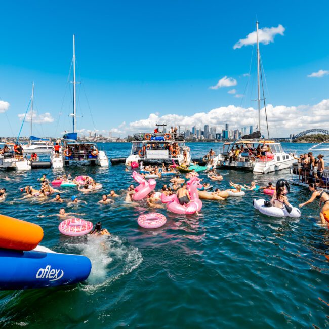 A vibrant scene of people enjoying a sunny day on the water, surrounded by sailboats and yachts. Many are floating on pink inflatable rings and large colorful rafts. The blue sky and distant city skyline add to the lively atmosphere, epitomizing the essence of The Yacht Social Club Sydney Boat Hire.