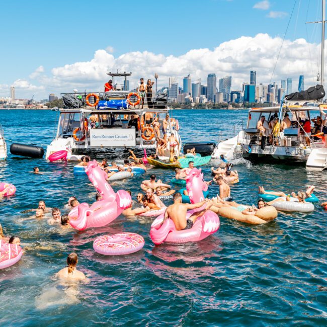 People are floating on large, colorful inflatable rafts shaped like pink flamingos in blue water, surrounded by boats. The skyline of a city with tall buildings is visible in the background under a bright blue sky. The scene, part of The Yacht Social Club's luxury yacht rentals in Sydney, is lively and festive.