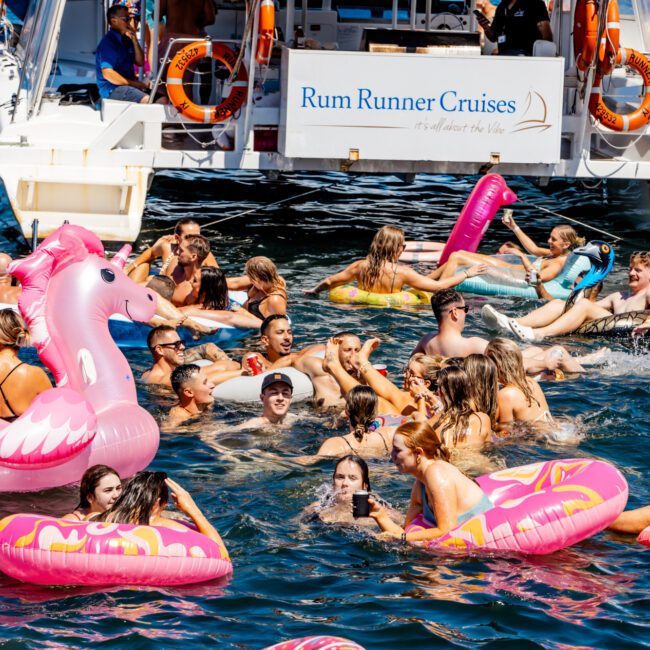 A lively pool party scene with people enjoying the water on various inflatable rafts, including pink flamingos. Behind them is a luxury yacht labeled "Rum Runner Cruises," courtesy of The Yacht Social Club Sydney Boat Hire. The sea is blue, and the atmosphere is festive and fun.