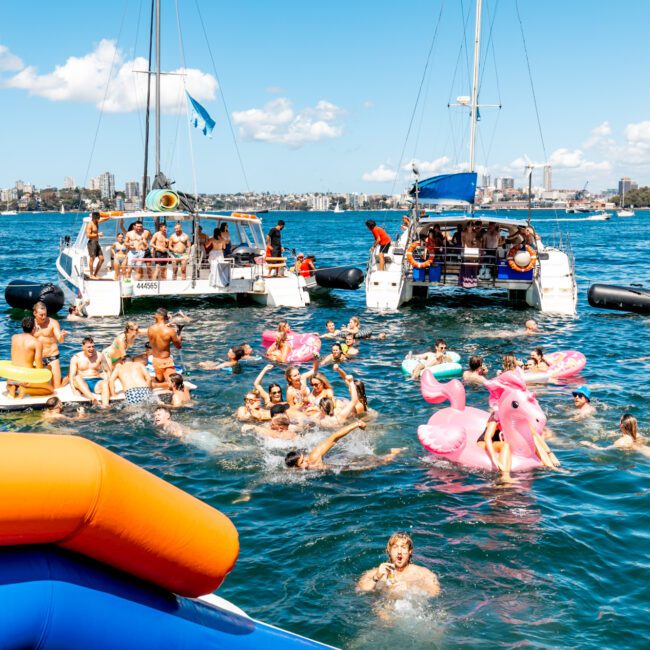 A vibrant scene of people enjoying a sunny day on the water, swimming and floating on colorful inflatables next to anchored boats. The Yacht Social Club Event Boat Charters animate the lively atmosphere as revelers make the most of Sydney Harbour. In the backdrop, a city skyline stretches under a blue sky with scattered clouds.