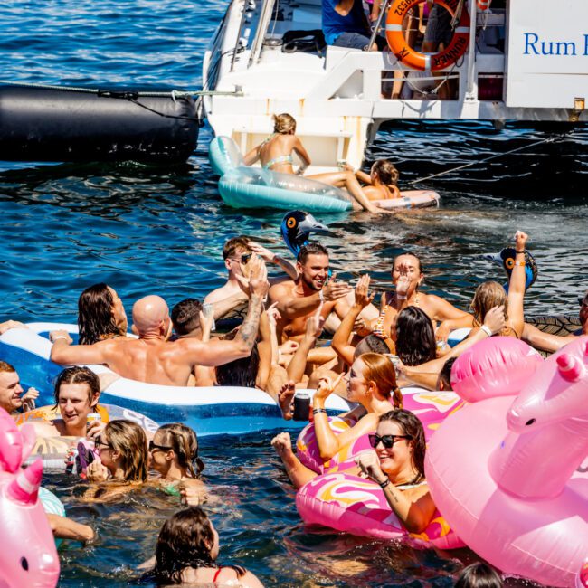 A lively scene of people having fun in the water near a boat. Many are using inflatable pink unicorn floats and rubber rings. Some are on a large floating mat, while others relax on and around the boat from Sydney Harbour Boat Hire The Yacht Social Club. The sky is clear and the atmosphere is festive.