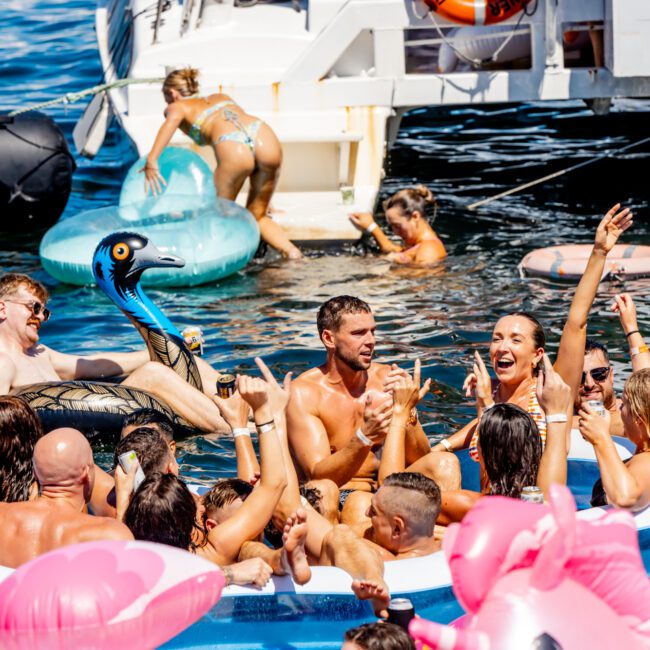 A large group of people enjoy a sunny day on the water, lounging on inflatable toys and a boat from The Yacht Social Club Sydney Boat Hire. They laugh, chat, and raise their hands in excitement as colorful inflatable animals float around the scene.