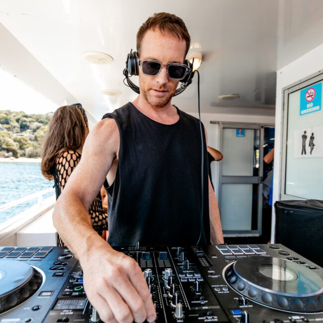 A man wearing sunglasses, a black sleeveless shirt, and a headset stands behind a DJ booth on a boat. He is adjusting the controls on the DJ equipment under bright sunlight. In the background, a woman enjoys the view of open water and shoreline. This scene captures The Yacht Social Club's vibrant Boat Parties Sydney.