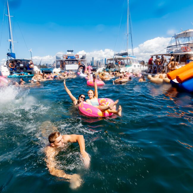 A lively party scene on the water features numerous people swimming and floating on various inflatable toys. Boats are anchored nearby, thanks to Luxury Yacht Rentals Sydney, and attendees are enjoying the sunny day with smiles and energetic activities. The sky is clear, and the atmosphere is festive.