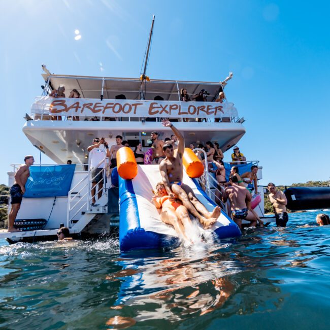 A group of people enjoys a sunny day aboard the "Barefoot Explorer" boat. Some individuals are sliding down an inflatable slide into the water, while others watch or swim nearby. The scene is lively, with clear blue skies and a surrounding view of nature and other boats. This fun-filled day is part of The Yacht Social Club Event Boat Charters, capturing the essence of luxury on the water.