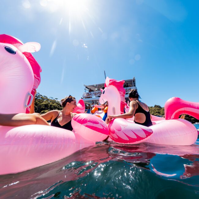 A group of people in swimwear are floating on large, inflatable pink unicorns in a body of water. They smile and chat under a sunny blue sky. In the background, there’s a boat from The Yacht Social Club Sydney Boat Hire and some trees. It’s a perfect day for boat parties with friends.