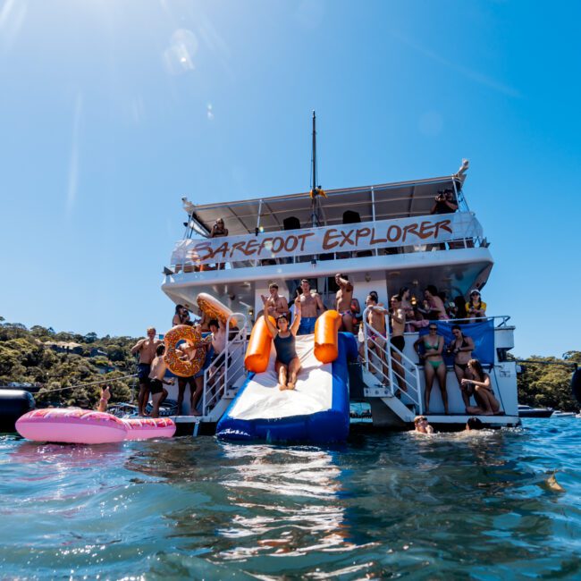 A group of people enjoy a sunny day on the "Barefoot Explorer" boat, which features a slide into the water. Several people are swimming and lounging on large pink and blue floats in the ocean. The background shows lush, green trees under a clear, blue sky. Perfect for Boat Parties Sydney The Yacht Social Club offers.