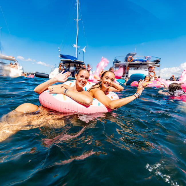 Two women are floating in the ocean on pink inflatable rings, smiling and waving at the camera. Other people are enjoying the water near luxurious yachts from Sydney Harbour Boat Hire. The festive atmosphere is enhanced by nearby boats moored under a clear blue sky.