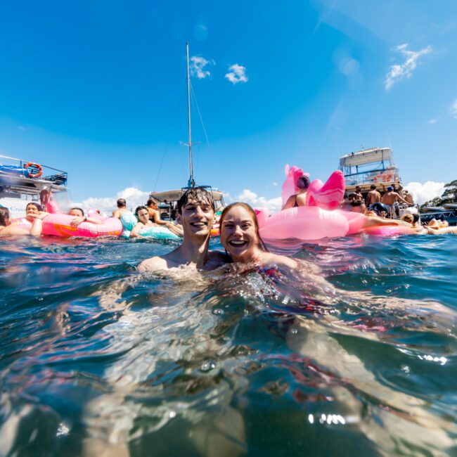 A group of people enjoying a sunny day in the water, some on inflatable floats. In the foreground, a smiling couple stands chest-deep in the water. In the background, boats from The Yacht Social Club Sydney Boat Hire and more people are participating in the fun. The sky is bright blue with some clouds.