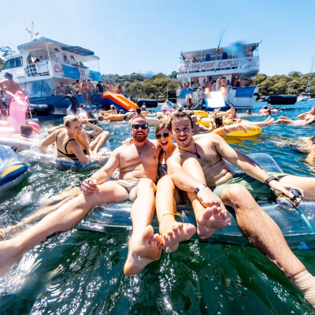 Group of people enjoying a sunny day on a lake, floating on inflatable pool toys, with boats in the background. Two men in the foreground cheerfully pose, lying on a transparent float. The scene is lively with laughter and bright colors from the floats and swimwear, reminiscent of The Yacht Social Club Sydney Boat Hire events.