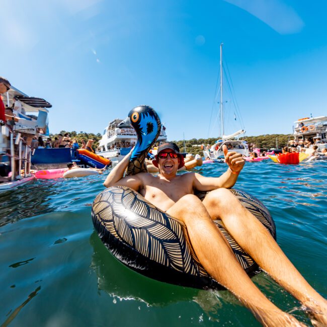 A person floats on a black, patterned inflatable ring in clear blue water, surrounded by boats and others enjoying a sunny day at Sydney Harbour Boat Hire. The individual holds an inflatable octopus while smiling and wearing sunglasses. The background includes a sailboat and a forested shoreline.