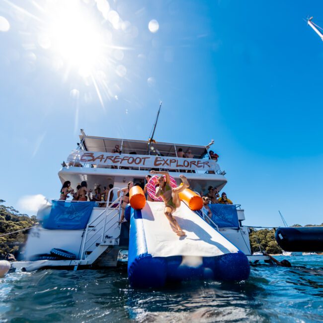 A girl in a yellow swimsuit slides down an inflatable slide from a boat named "Barefoot Explorer" into the water. Other people are on the boat and in the water, enjoying the sunny day with clear blue skies. Trees are visible in the background, creating an idyllic scene reminiscent of The Yacht Social Club's luxury yacht rentals in Sydney.