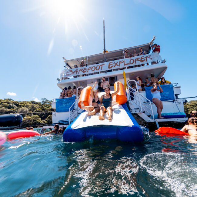 A vibrant scene featuring people enjoying a sunny day on a boat named "Barefoot Explorer," hosted by The Yacht Social Club Sydney Boat Hire. Individuals are sliding down an inflatable slide into the clear blue water, while others relax on colorful floaties. The sky is bright blue, and the mood is cheerful and lively.