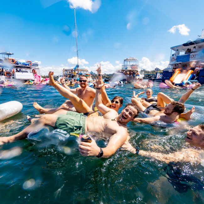 A group of people enjoying a sunny day on the water, floating on inflatables, and laughing. Boats are anchored nearby, showcasing Sydney Harbour Boat Hire The Yacht Social Club. The background shows a clear blue sky with fluffy clouds. The scene is lively and festive with people engaging in summertime fun.