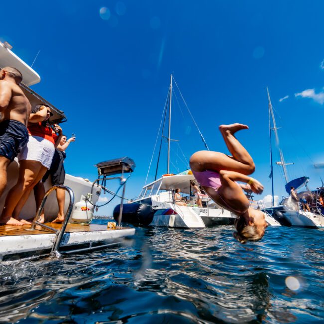 A person in a swimsuit performs a backflip into the water from a boat as people on the deck watch. Several boats are anchored nearby under a clear blue sky, enjoying The Yacht Social Club Event.