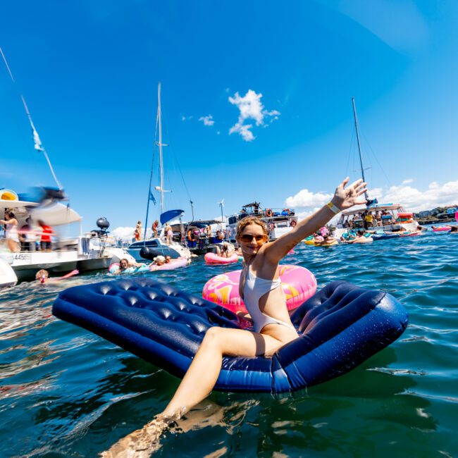 A person in a swimsuit lies on a blue inflatable mattress in a sunny, crowded marina. They are waving towards the camera and smiling, surrounded by boats and other floaties in the water. The sky is clear with few clouds, adding to the vibrant ambiance of The Yacht Social Club Sydney Boat Hire.