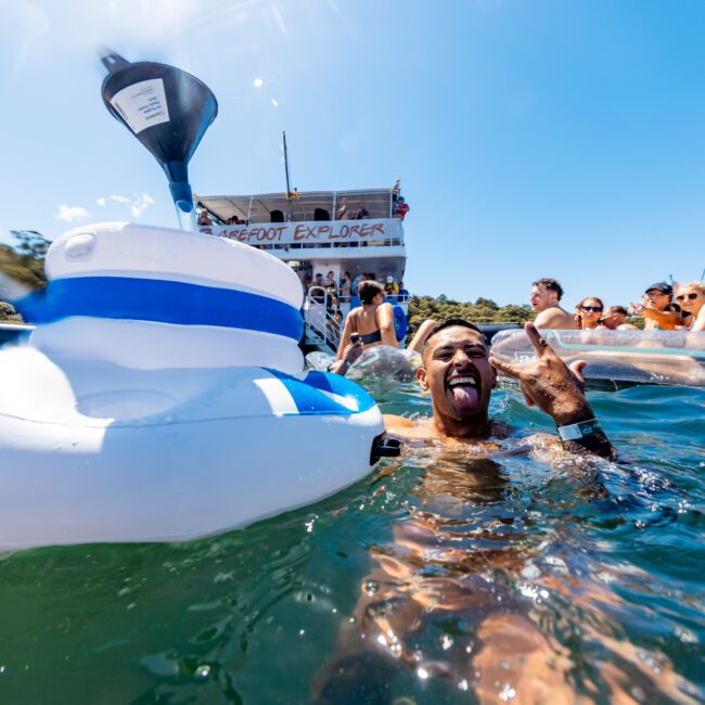 A group of people are enjoying a sunny day on a boat named "Barefoot Explorer." Some are in the water, including a man making a playful face and hand gesture near an inflatable toy. Other boats and scenic hills are visible in the background, showcasing The Yacht Social Club Event Boat Charters.