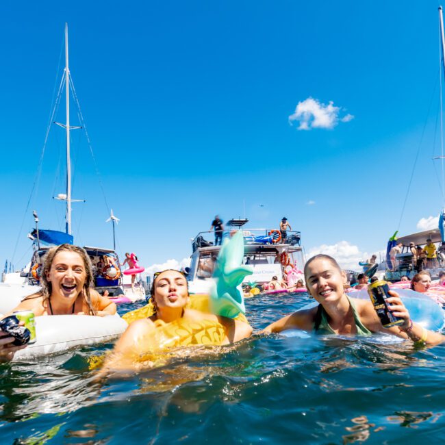 Three people are enjoying a sunny day on the water, smiling and floating with boats and other people in the background. One person holds a drink, and they all appear to be having a fun and lively time. The sky is clear with a few clouds visible—a perfect scene for Luxury Yacht Rentals Sydney.