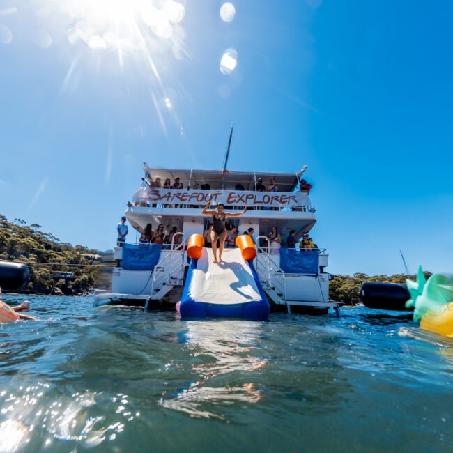 A vibrant scene of a boat named "Barefoot Explorer" in clear blue water. People are enjoying the sun and water slide off the back of the boat. Various colorful inflatables, including a pineapple and a unicorn, float around while lush greenery sets the backdrop. Perfect for a The Yacht Social Club Sydney Boat Hire experience.