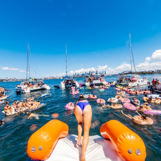 A person in a blue swimsuit stands at the edge of a boat, preparing to jump into the water. The scene is lively, with numerous people on nearby boats and floatation devices enjoying the sunny day on the lake. A clear blue sky and distant city skyline add to the charm of The Yacht Social Club Event Boat Charters.