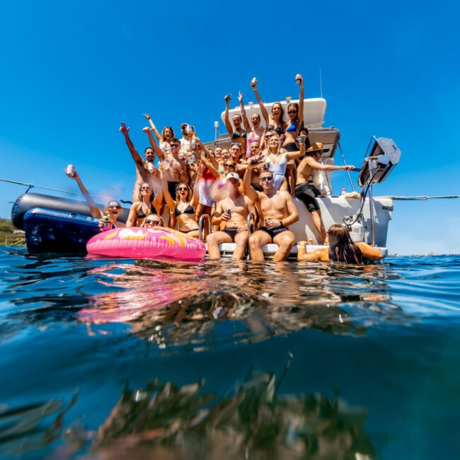 A large group of people are posing and cheering on the back of a boat from Luxury Yacht Rentals Sydney. The clear blue sky and calm water suggest a sunny day. Some individuals are in the water, near a pink inflatable ring. The mood is lively and festive, perfect for Boat Rental and Parties Sydney The Yacht Social Club.