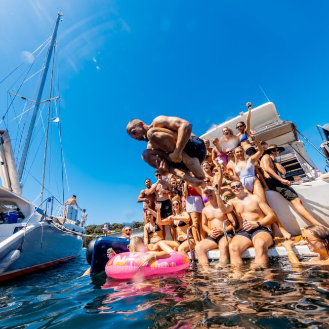 A group is enjoying a sunny day on a boat rental from Sydney Harbour Boat Hire The Yacht Social Club. Some are cheering on the deck, while others splash in the water. A person mid-air is about to cannonball, and children play on a pink inflatable in the foreground.