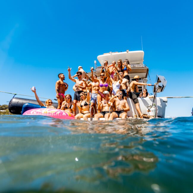 A group of people in swimwear are gathered on the back of a boat from The Yacht Social Club, enjoying a sunny day. Some are sitting on the edge, others are standing, and a few are in the water. A bird flies in the clear blue sky above them.