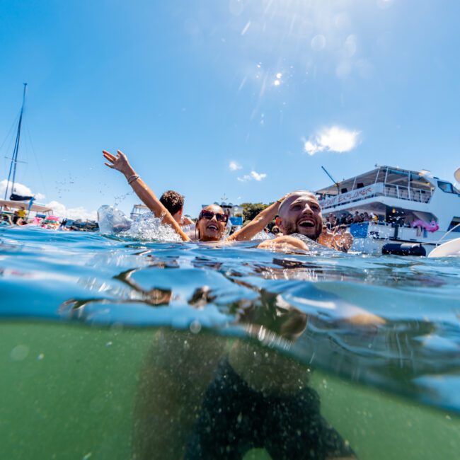 Two smiling people are floating in the water, with one cheerfully raising their arms. The photo is partly underwater and shows a yacht from The Yacht Social Club in the background under a clear blue sky. The light creates a vibrant, joyful atmosphere on Sydney Harbour.