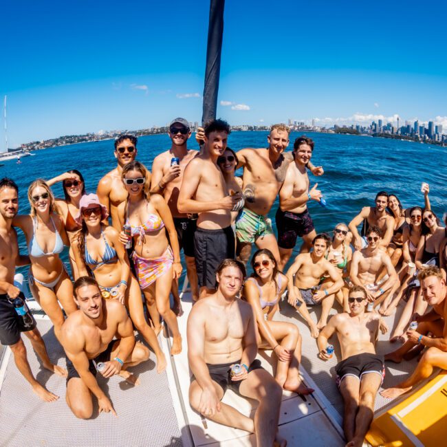 A large group of young adults in swimwear are gathered on a sailboat, enjoying a sunny day at sea. The backdrop features a clear blue sky, the city skyline, and numerous sailboats on the water. It's evident they are attending The Yacht Social Club Sydney Boat Hire event, smiling and socializing under the sun.