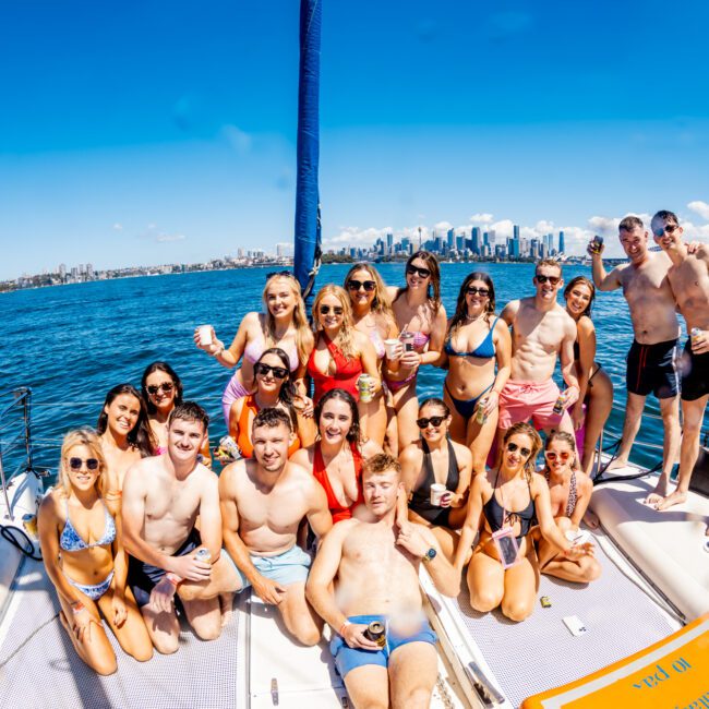 A group of people are on a boat, posing for a photo under a clear blue sky. They are all in swimwear, smiling, and holding drinks. The city skyline and other boats are visible in the background. They appear to be enjoying a sunny day on the water at The Yacht Social Club event hosted by Luxury Yacht Rentals Sydney.