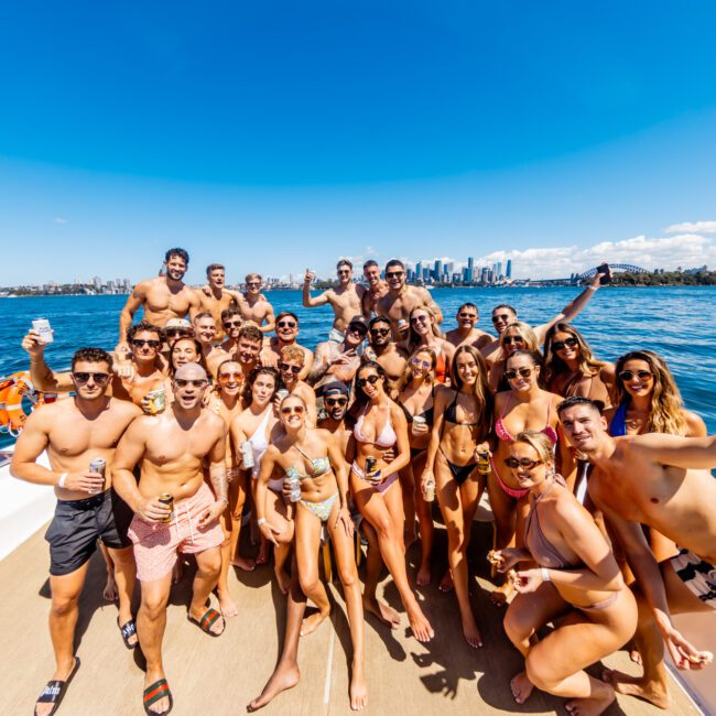 A large group of people in swimwear are gathered on a boat, smiling and posing for the camera. They are surrounded by water with a city skyline visible in the background under a clear blue sky, capturing the vibrant spirit of The Yacht Social Club Event Boat Charters.