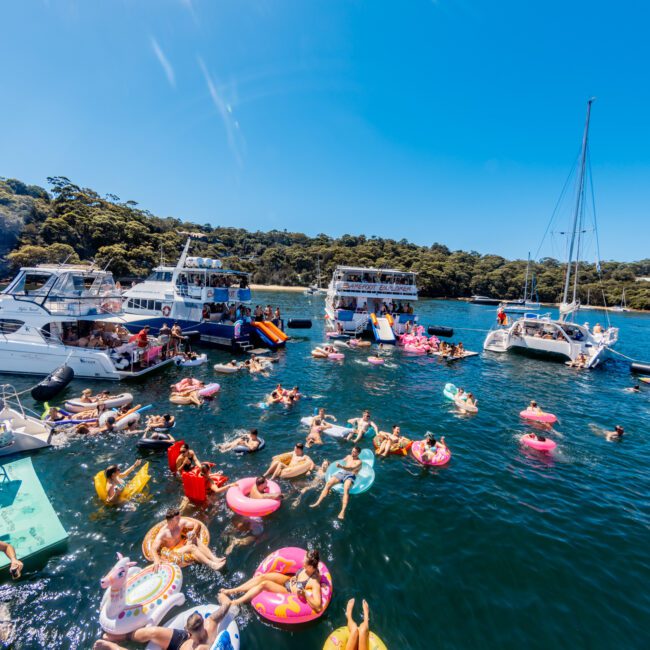 People enjoying a sunny day on the water, swimming and lounging on various colorful inflatables like rings and mats, surrounded by anchored boats and yachts. The Yacht Social Club Event Boat Charters add an extra layer of excitement to this vibrant scene, all against lush greenery under a clear blue sky.