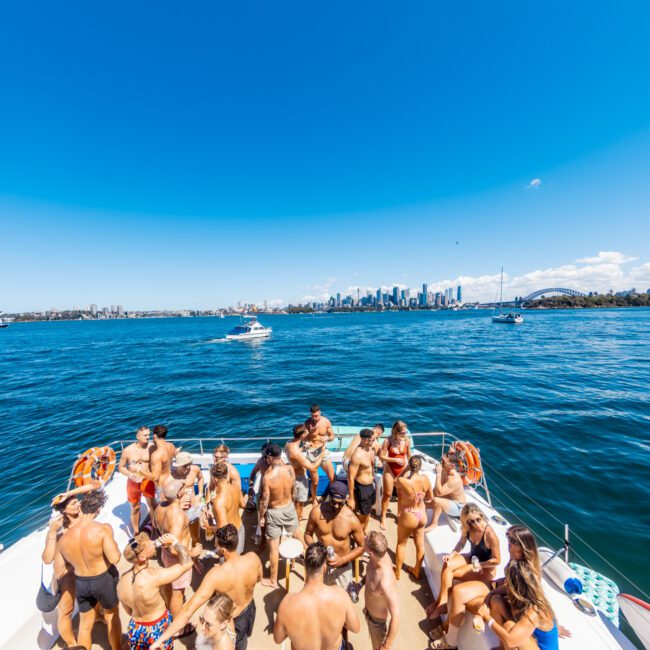 A group of people in swimwear enjoying a sunny day on a boat from Sydney Harbour Boat Hire The Yacht Social Club. The boat is on a large body of water with a city skyline and a bridge visible in the background. The sky is clear and blue, with a few scattered clouds.