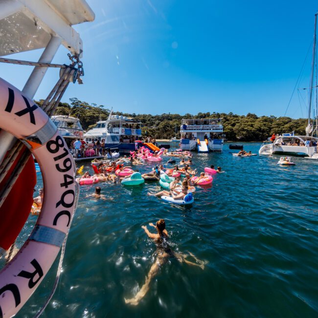 A group of people swimming and floating on colorful inflatables in a sunny blue water bay, surrounded by several anchored boats. A life preserver with visible numbers is in the foreground, and a green, tree-lined shore is in the background. Enjoy the ultimate experience with Boat Parties Sydney The Yacht Social Club.