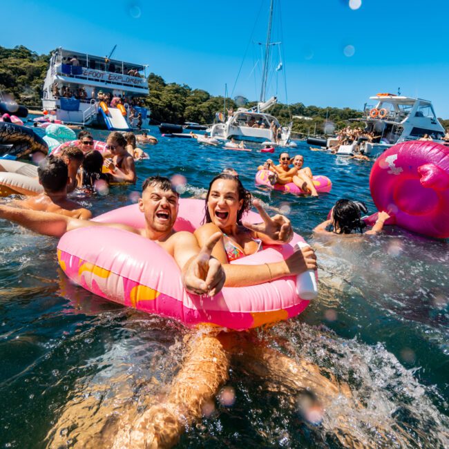 A joyful man and woman float in pink donut-shaped pool inflatables, smiling and giving thumbs up. Surrounded by other people on inflatables and boats from The Yacht Social Club Sydney Boat Hire, they enjoy a sunny, vibrant lake or ocean setting. Fun atmosphere with blue skies and lush greenery in the background.