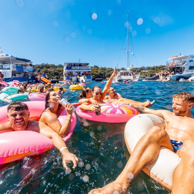 A group of people enjoys a sunny day in the water with inflatables, surrounded by boats. Some are drinking and others relaxing on floating devices, all part of The Yacht Social Club Event Boat Charters. The background shows a clear blue sky and a forested shoreline.